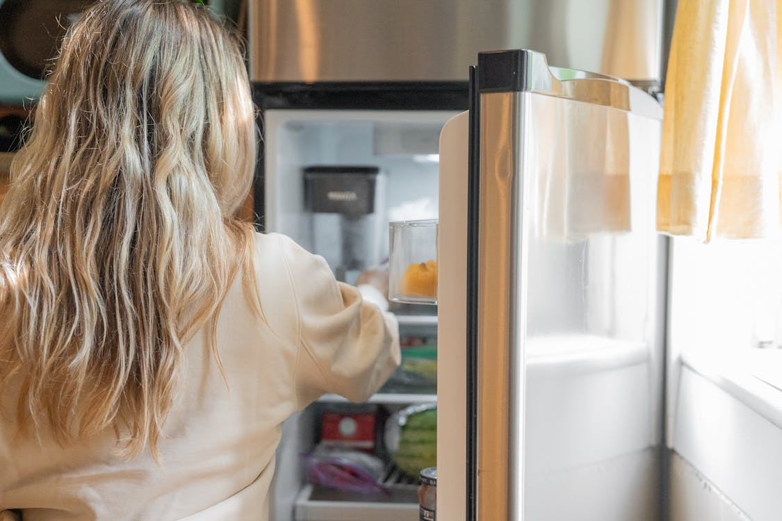 The image shows a person with wavy blonde hair reaching into a refrigerator.