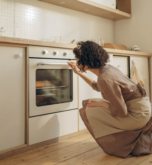 The image shows a person kneeling and checking something inside an oven in a modern kitchen.